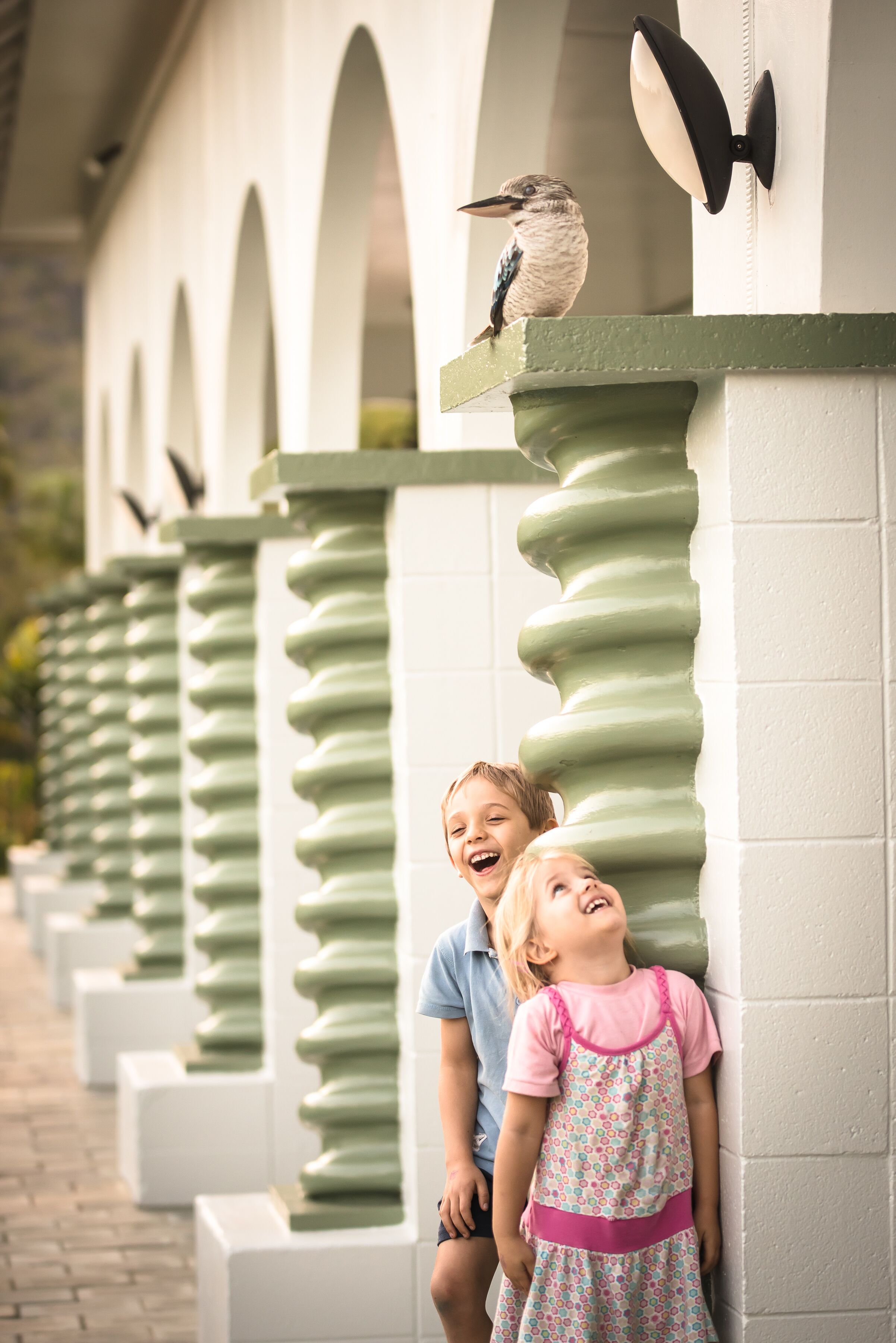 A child and an adult leaning against a building with green columns and a bird perched on a ledge.