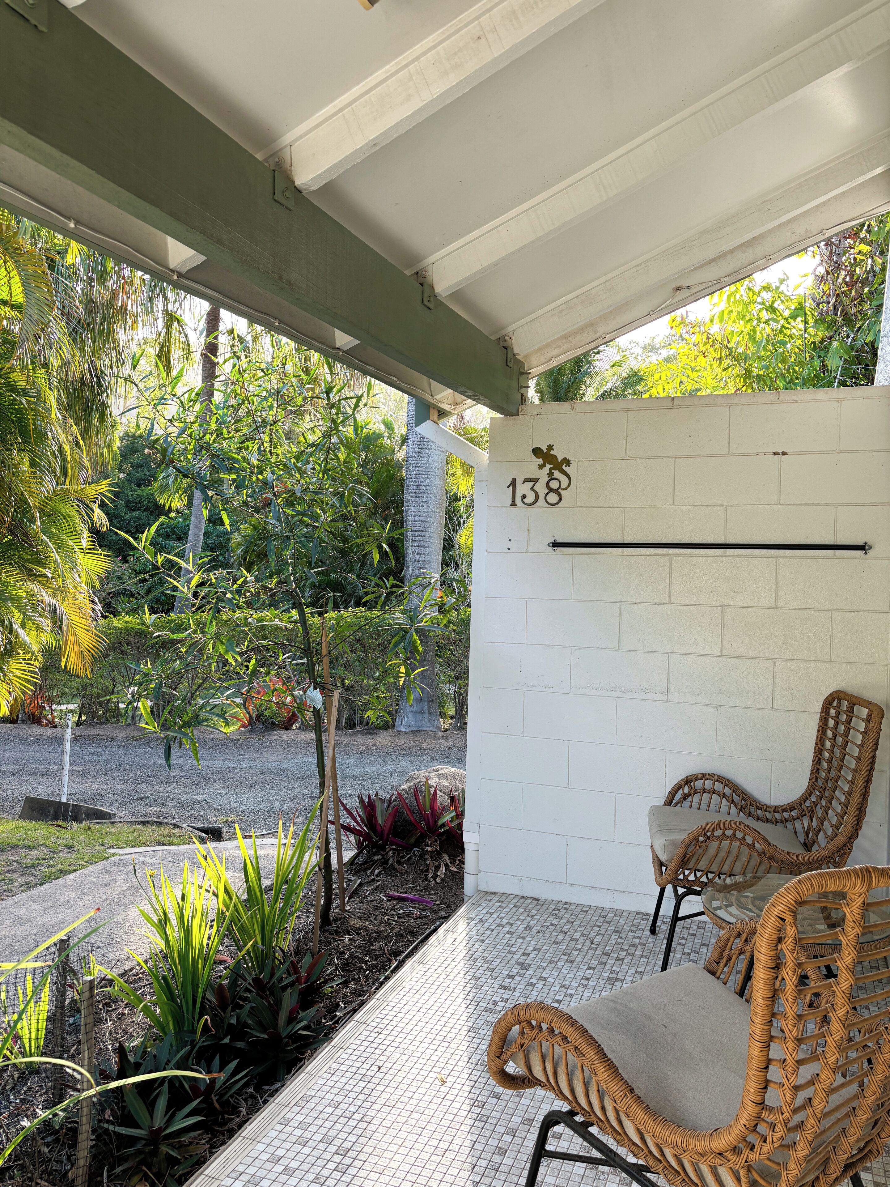 A covered outdoor seating area with wicker chairs, a white brick wall with the number 138, and a view of a garden with various plants and trees.