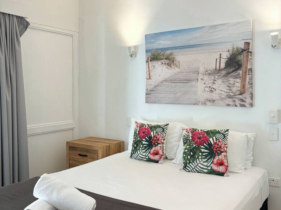 A neatly made bed with a dark brown bedspread, two pillows with tropical floral patterns, and a rolled white towel on the bed.