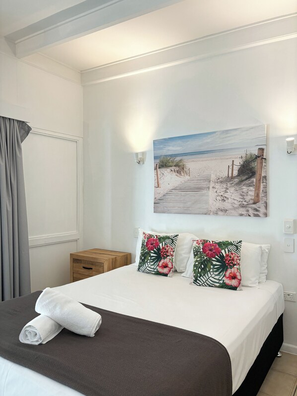 A neatly made bed with a dark brown bedspread, two pillows with tropical floral patterns, and a rolled white towel on the bed.