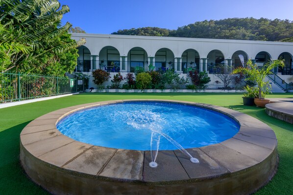 A swimming pool with water fountains, surrounded by a green lawn and a building with arched windows in the background.