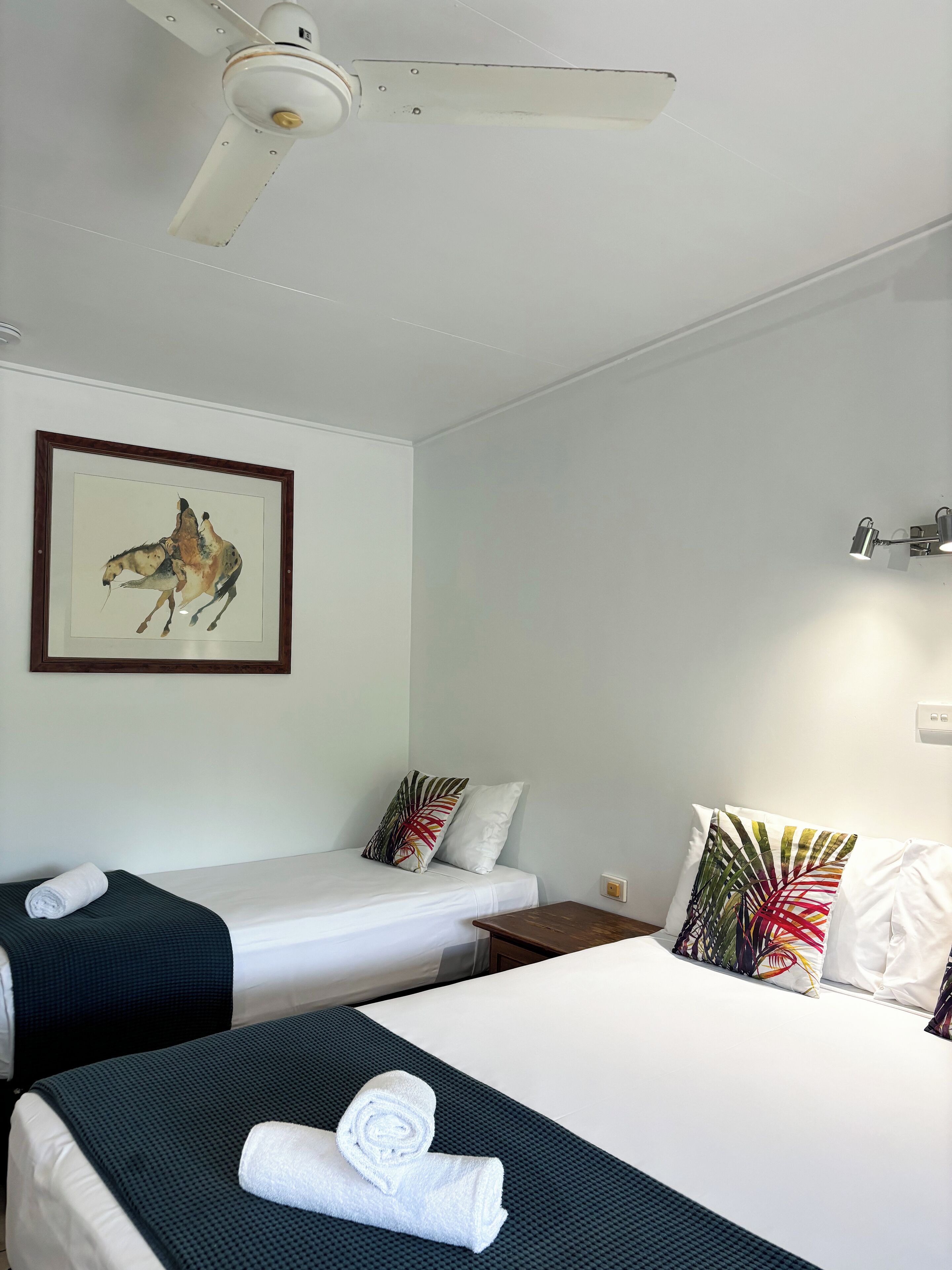 Habitación de hotel con dos camas individuales, un ventilador de techo, una obra de arte enmarcada de un caballo y una lámpara fijada a la pared.