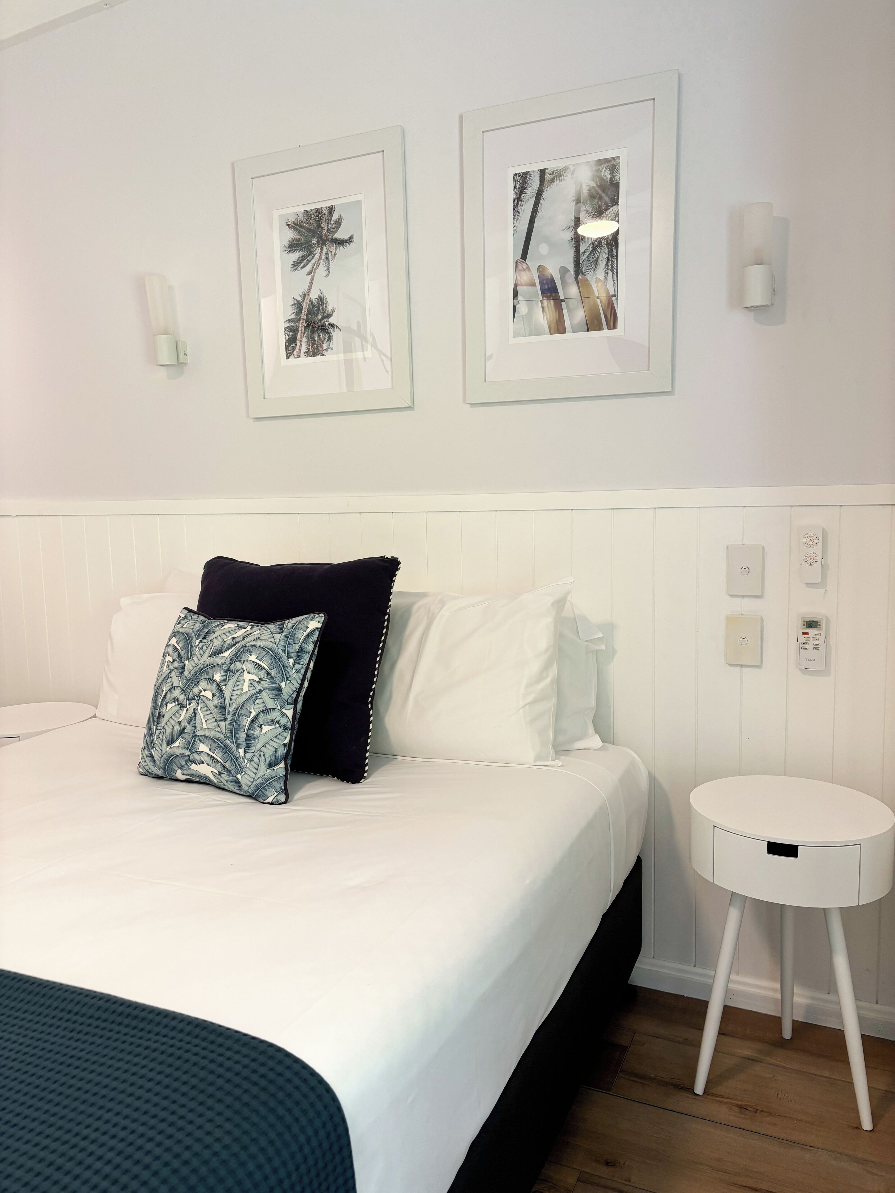 A hotel room with a bed, white walls, two framed pictures, a white bedside table, and a small white lamp.
