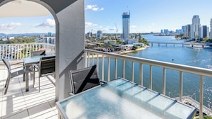 Un balcón con mesa y sillas de vidrio, con vista a un río y un paisaje urbano con un puente al fondo.