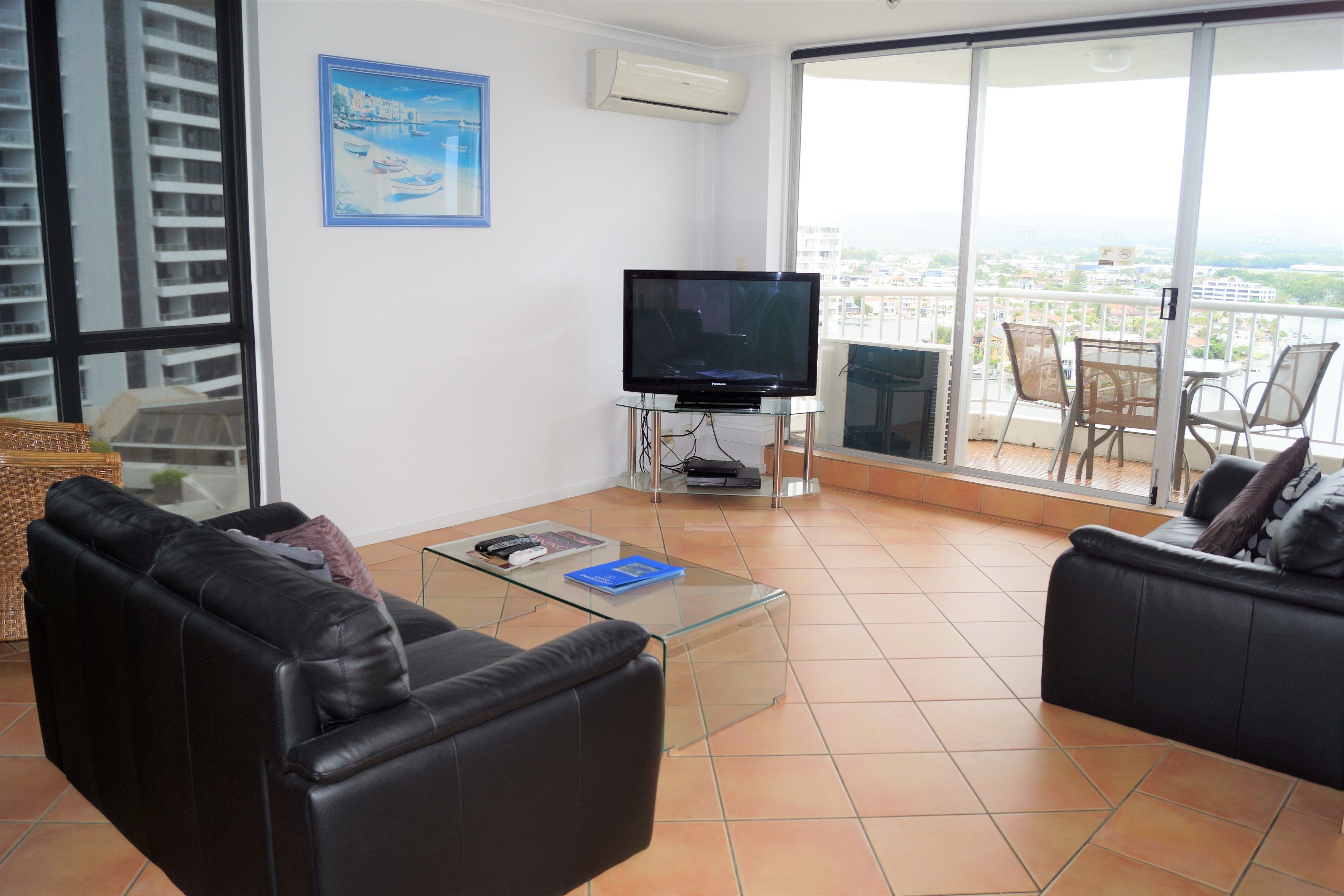 A living room with a glass coffee table, two black leather sofas, a flat-screen TV on a stand, and a large sliding glass door leading to a balcony with outdoor furniture.