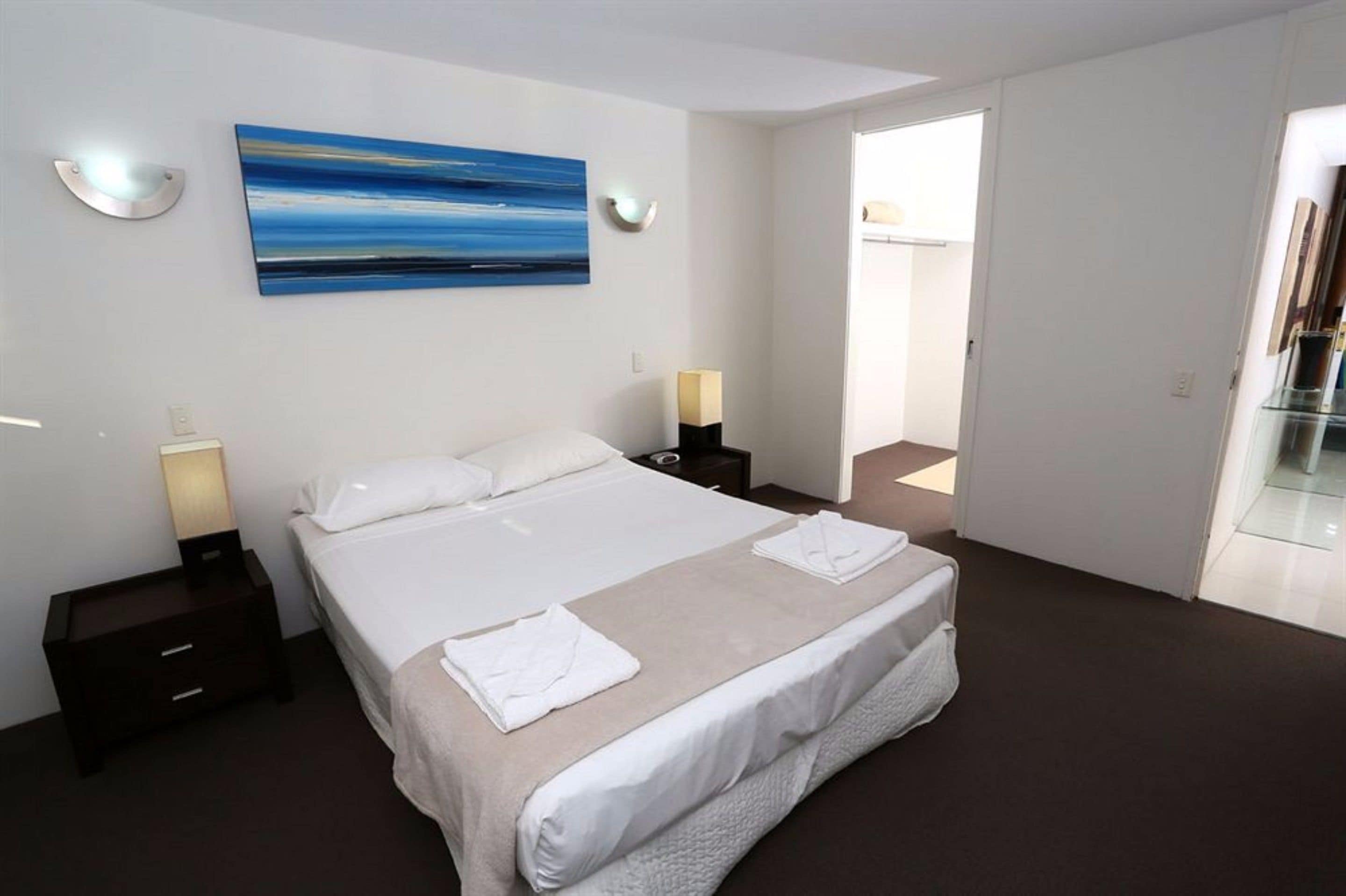 A modern hotel room with a large bed, two bedside tables with lamps, a painting on the wall, and a glass door leading to a balcony.