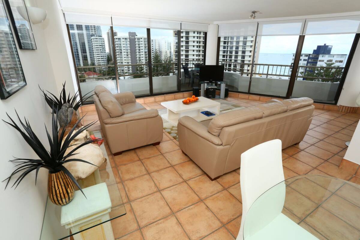 A spacious living room with a large sofa, a coffee table, and a TV. There is a balcony with a view of the city and the sea.