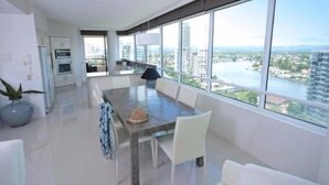 A modern living room with a large dining table, white furniture, and a view of a cityscape through large windows.