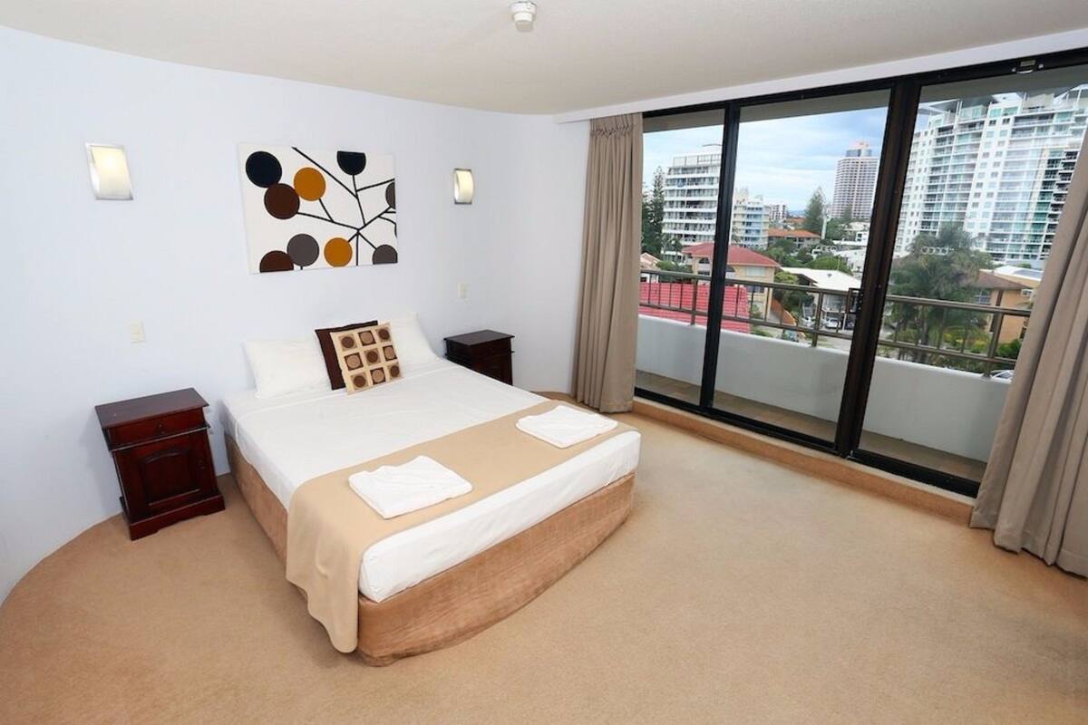 A hotel room with a large bed, a nightstand, a wall art piece, and a balcony with a city view.