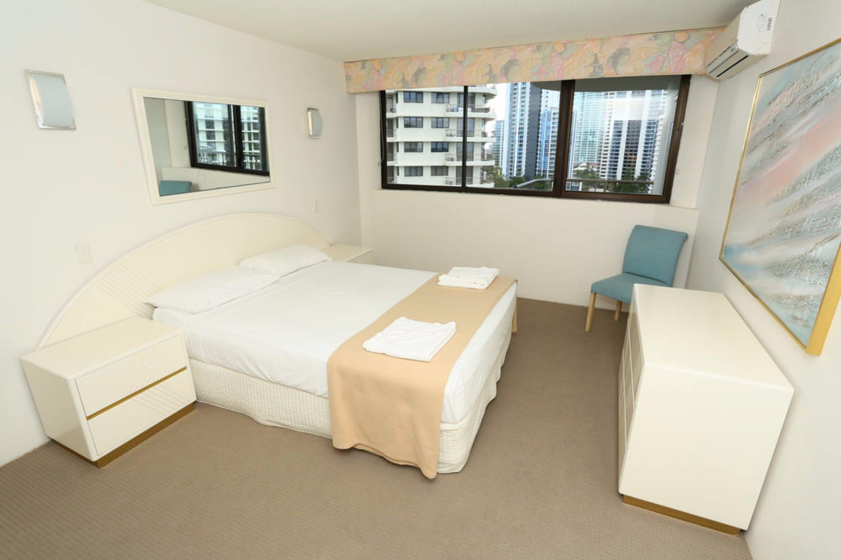 A hotel room with a bed, bedside tables, a desk, a chair, a large window, and a painting on the wall.