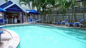 Una piscina con sillas de descanso y sombrillas, rodeada de palmeras y un edificio con techo azul.