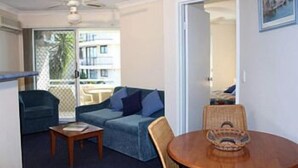 Una habitación con un sofá azul, una mesa de madera y una silla. Hay un balcón con vistas a edificios y una palmera.
