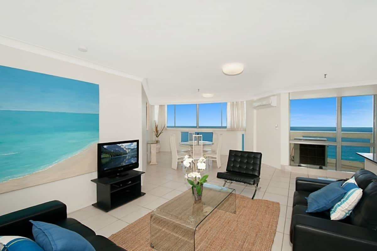 A modern living room with a large painting of a beach, a flat-screen TV, a dining area with a table and chairs, and a sofa with cushions.