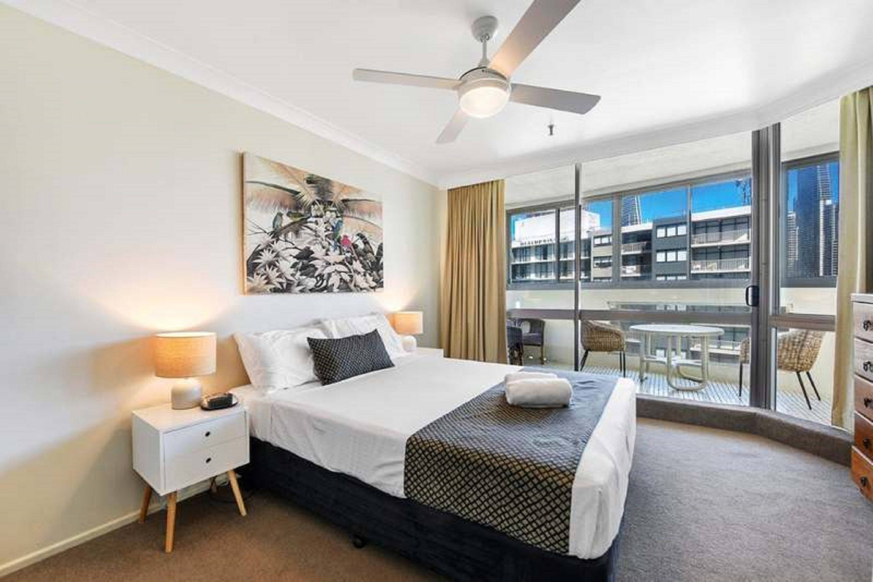 A hotel room with a bed, a ceiling fan, a large window, and a balcony with outdoor furniture.