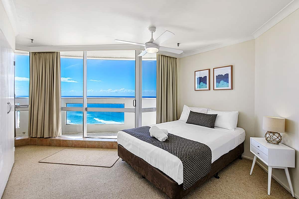 A hotel room with a large bed, a ceiling fan, a nightstand, and a view of the ocean through a sliding glass door.