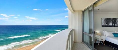 A balcony with a view of the ocean, a bed, and a small table with a lamp.