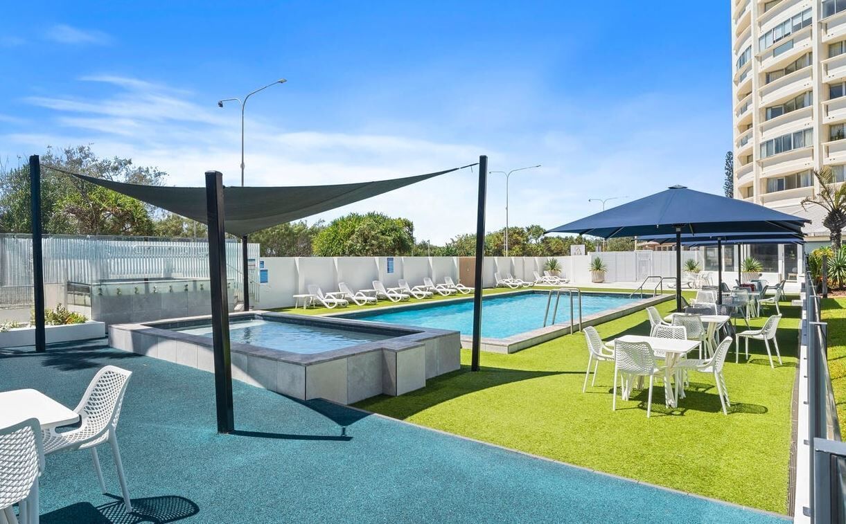 A rooftop pool area with lounge chairs and tables, surrounded by a low fence and a tall building in the background.