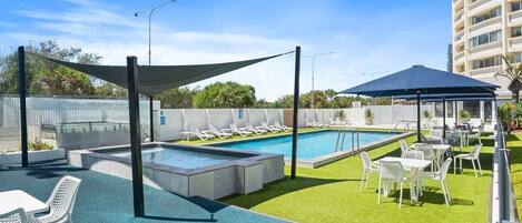 A rooftop pool area with lounge chairs and tables, surrounded by a low fence and a tall building in the background.