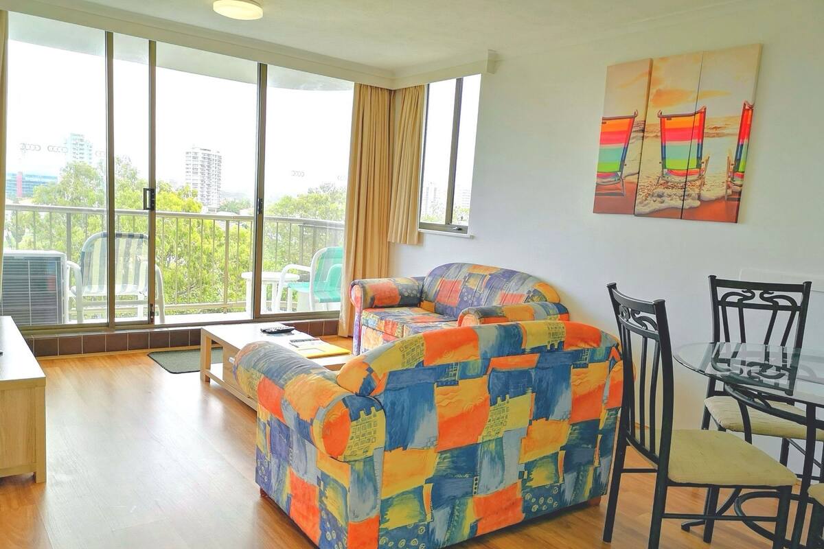 En moderne stue med en farverig sofa, et spisebord med stole og store vinduer med udsigt over byen.