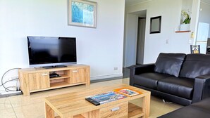 En moderne stue med et fladskærms-tv på et træstel, en sort lædersofa og et træsofabord med magasiner.