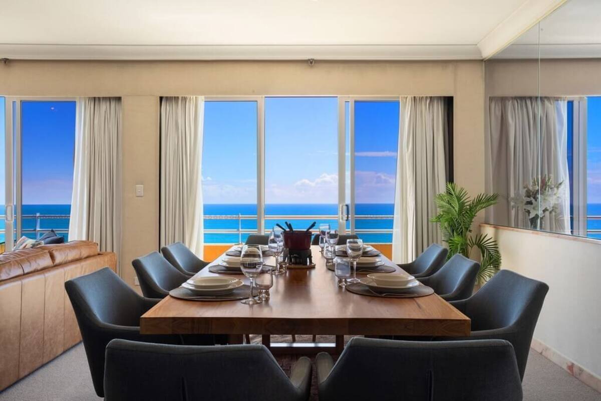 Un coin repas avec vue sur l’océan, une table en bois dressée pour quatre personnes et un canapé offrant également une vue imprenable.