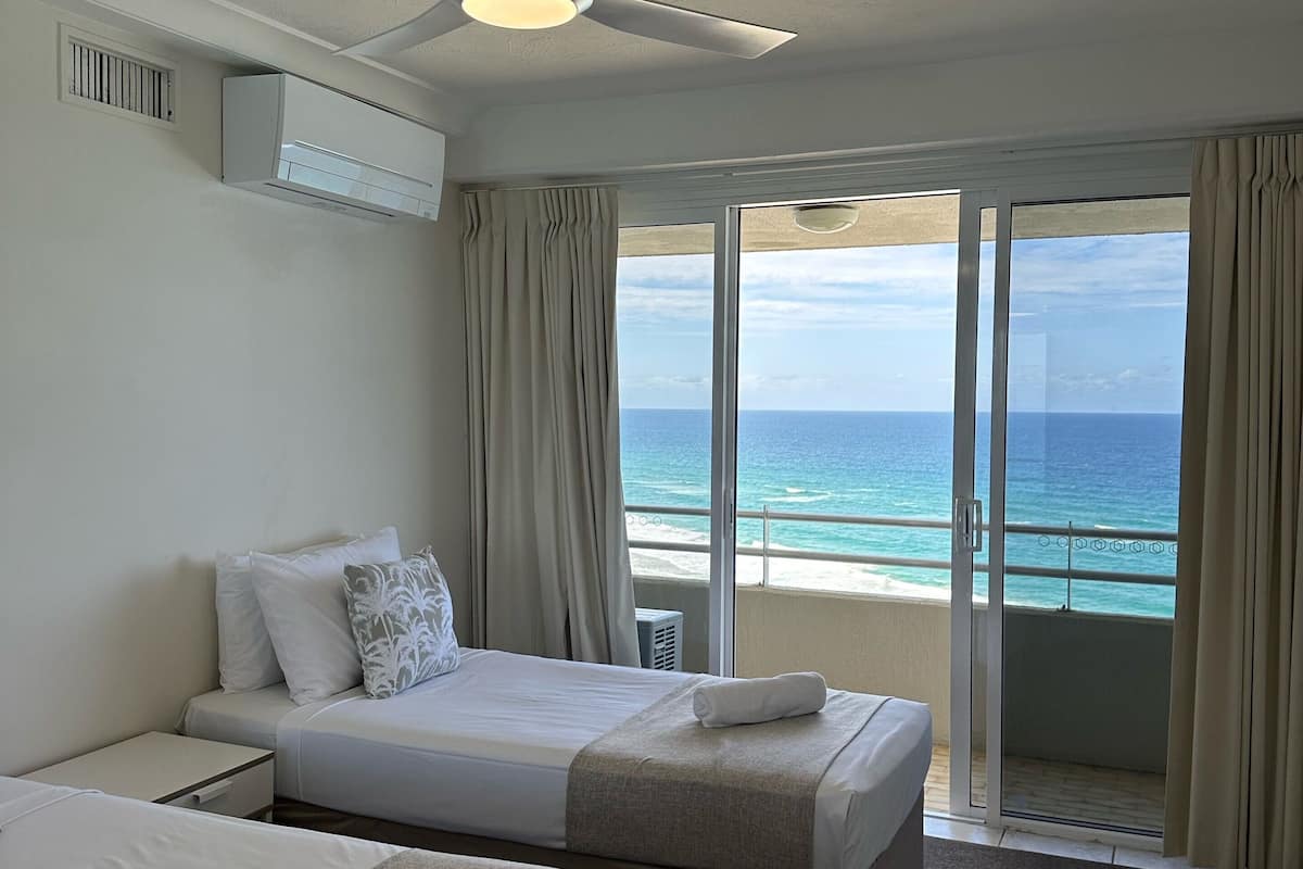 Une chambre d’hôtel avec deux lits, un ventilateur de plafond, un appareil de climatisation et une vue sur l’océan grâce à de grandes portes-fenêtres coulissantes.
