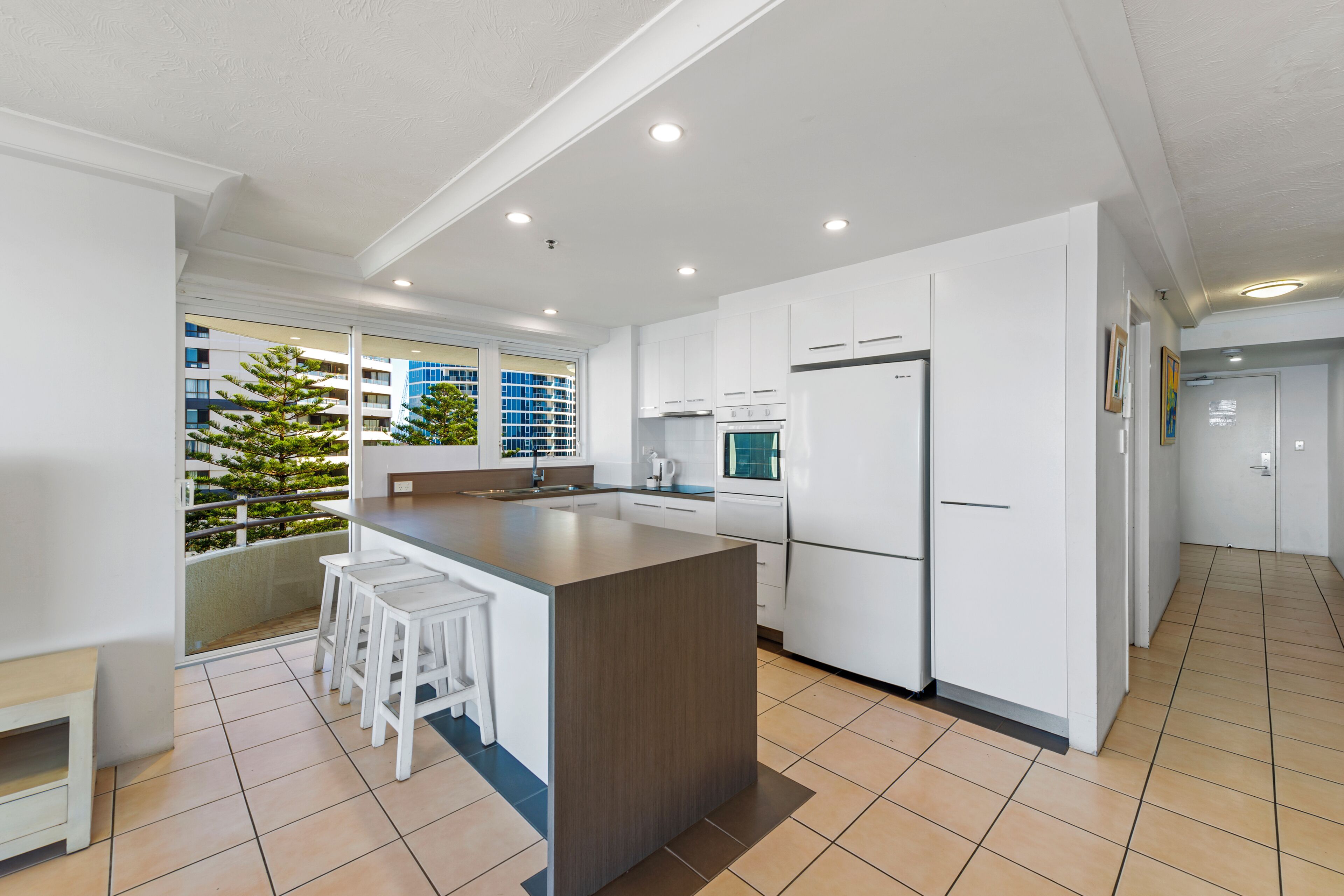A modern kitchen with white cabinets, a central island with stools, a built-in oven, and a refrigerator.
