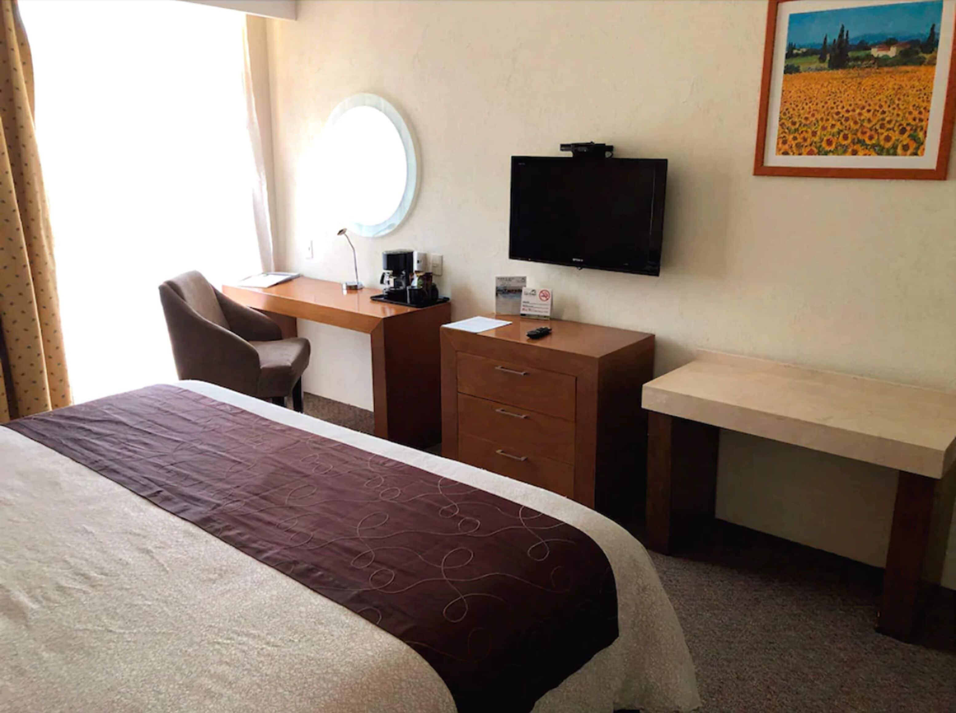 A hotel room with a bed, a desk, a chair, a television, and a painting on the wall.