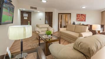 Ein Wohnzimmer mit einer Couch, einem Sessel und einem Couchtisch. An der Wand ist ein Fernseher angebracht und auf einem Beistelltisch steht eine Lampe.