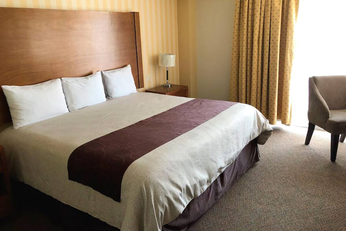 A hotel room with a large bed, a wooden headboard, a nightstand with a lamp, a chair, and a window with curtains.