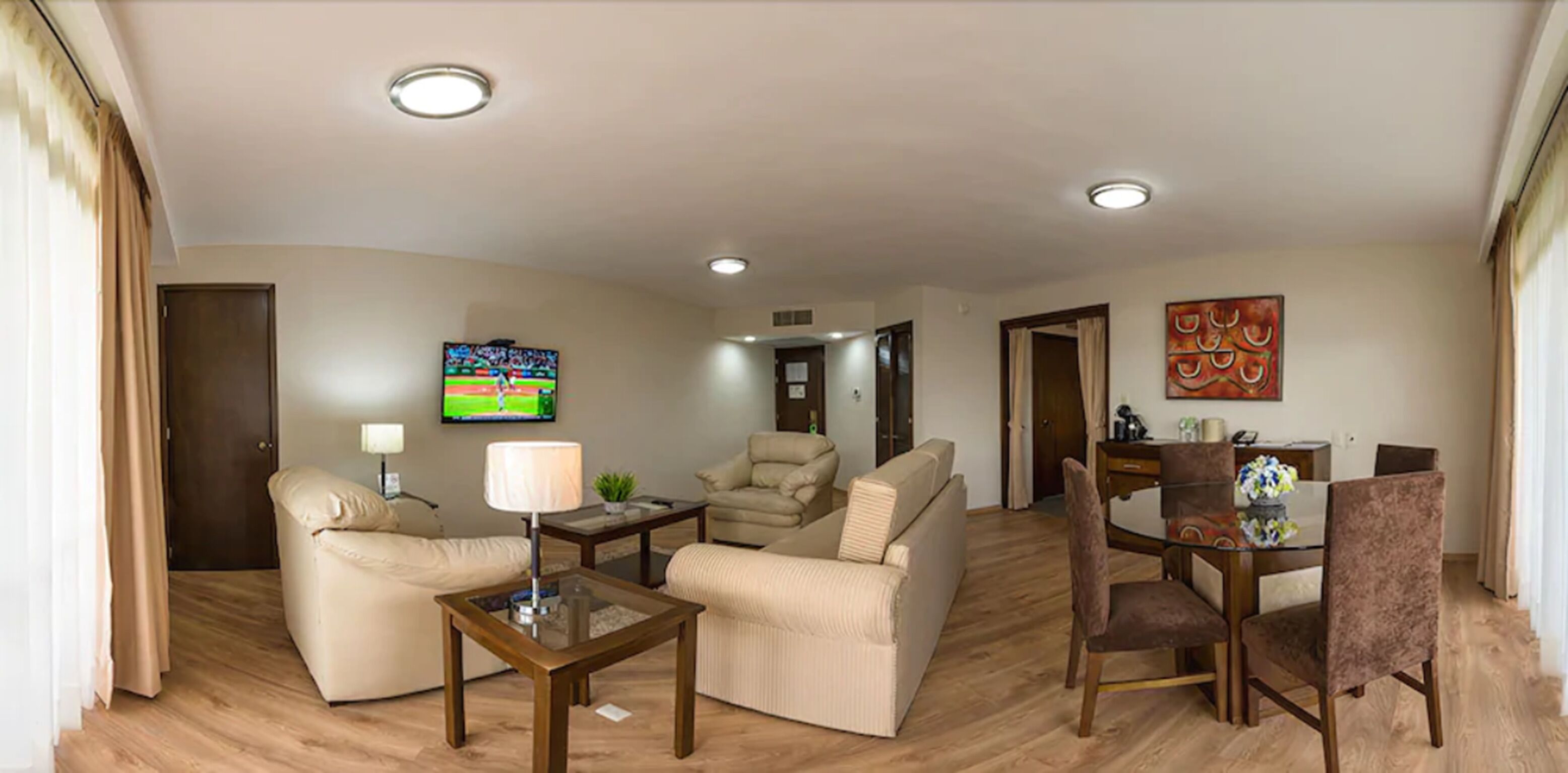 Una sala de estar moderna con un sofá, un sillón y una mesa de comedor con sillas. Hay un televisor montado en la pared y un cuadro sobre la mesa de comedor.