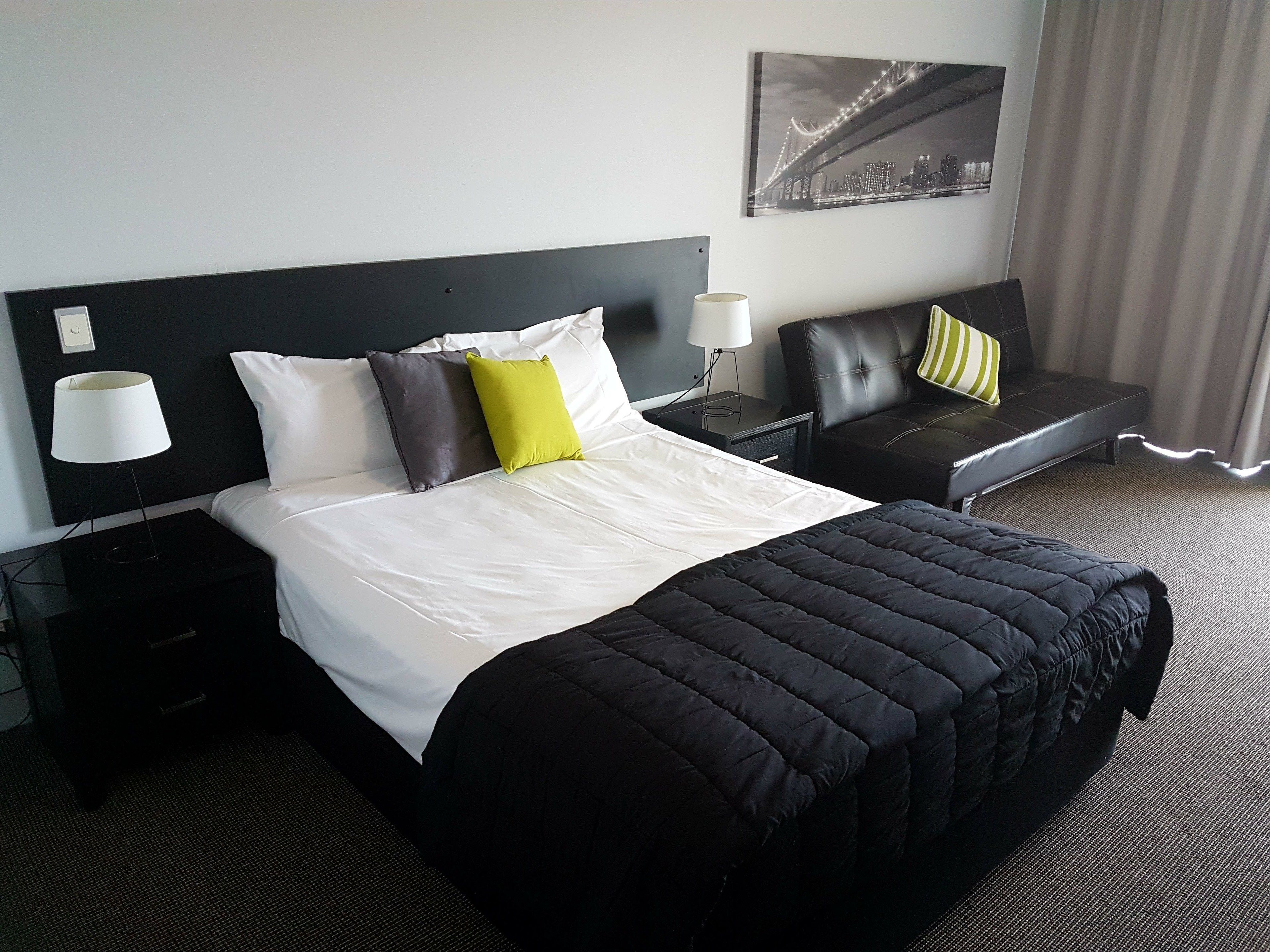 A hotel room with a large bed, a nightstand, a lamp, a chair, and a cityscape painting on the wall.