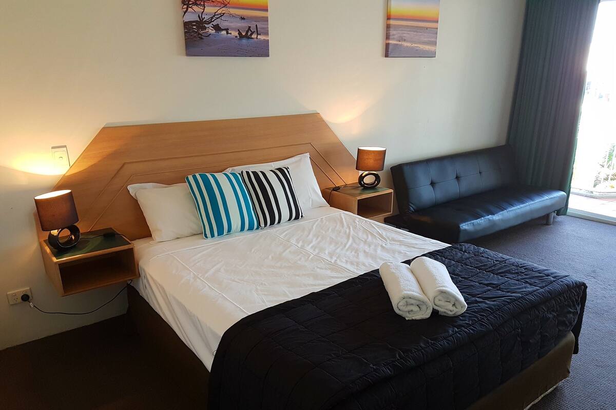 A hotel room with a bed, two bedside lamps, a nightstand, a black sofa, and two wall art pieces.