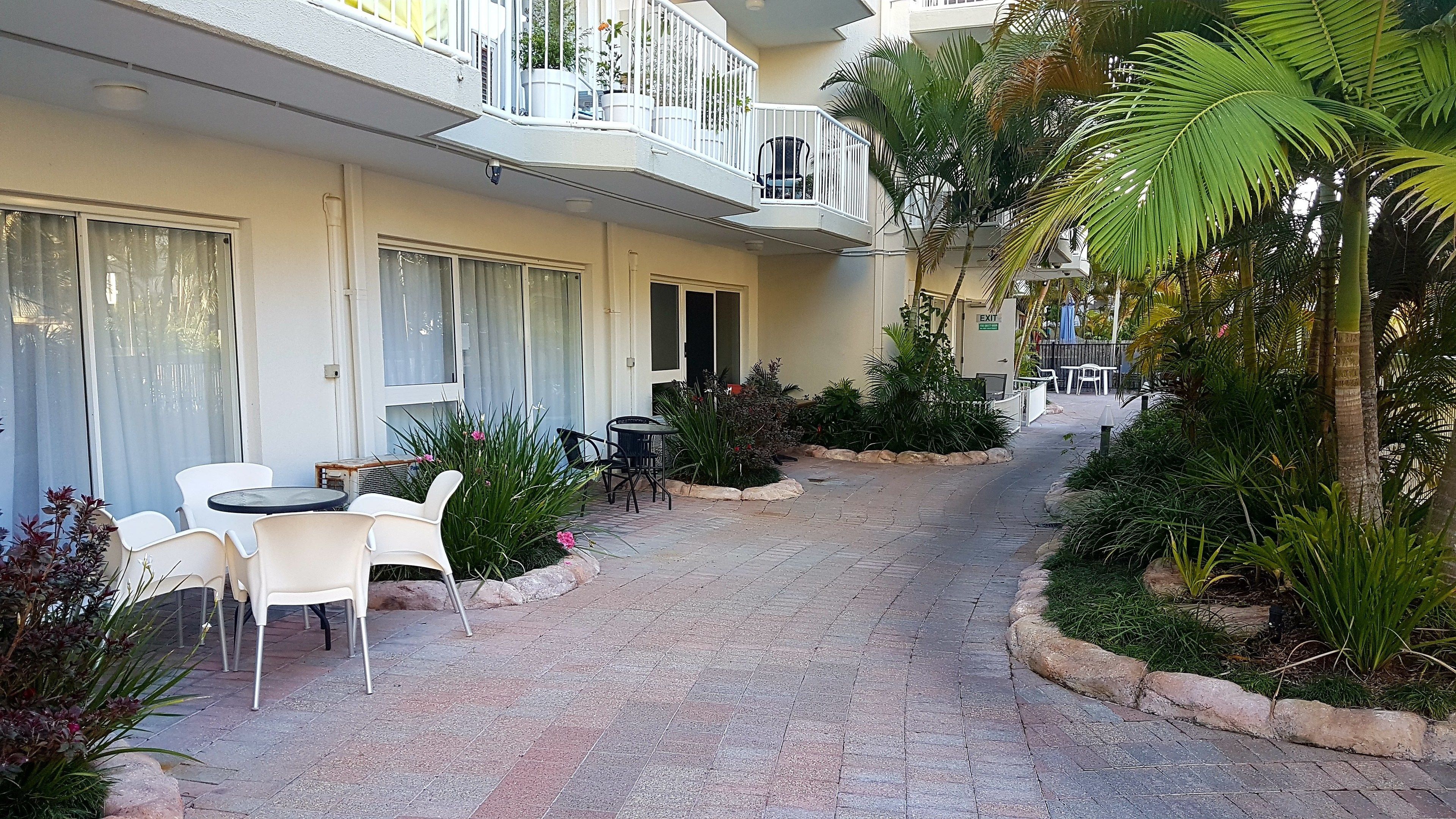 A hotel corridor with a paved walkway, white outdoor seating, and lush greenery.