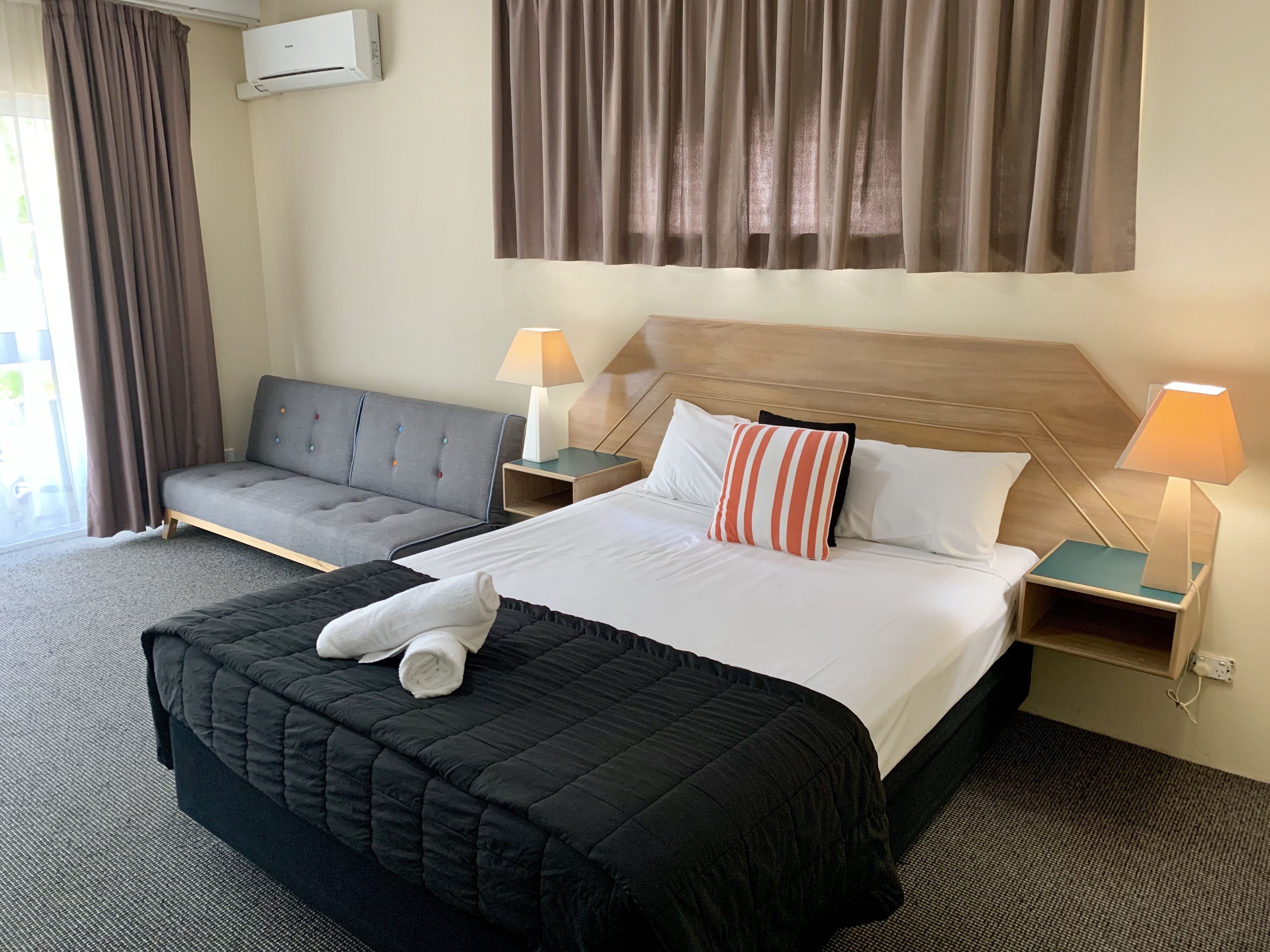 A hotel room with a bed, a sofa, two bedside lamps, a window with curtains, and an air conditioning unit.