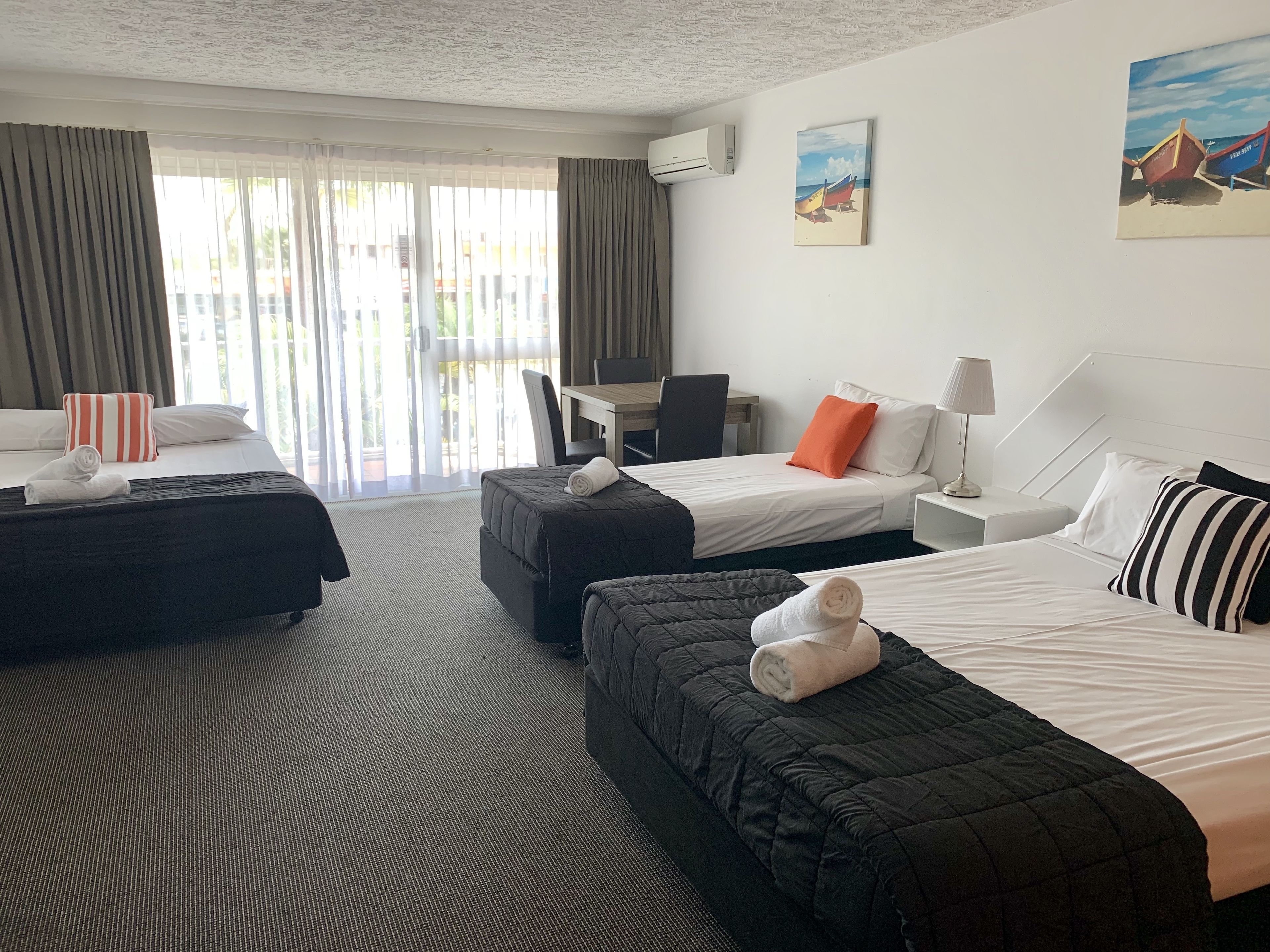 A hotel room with two beds, a desk, and a chair. There is a painting on the wall and an air conditioning unit.