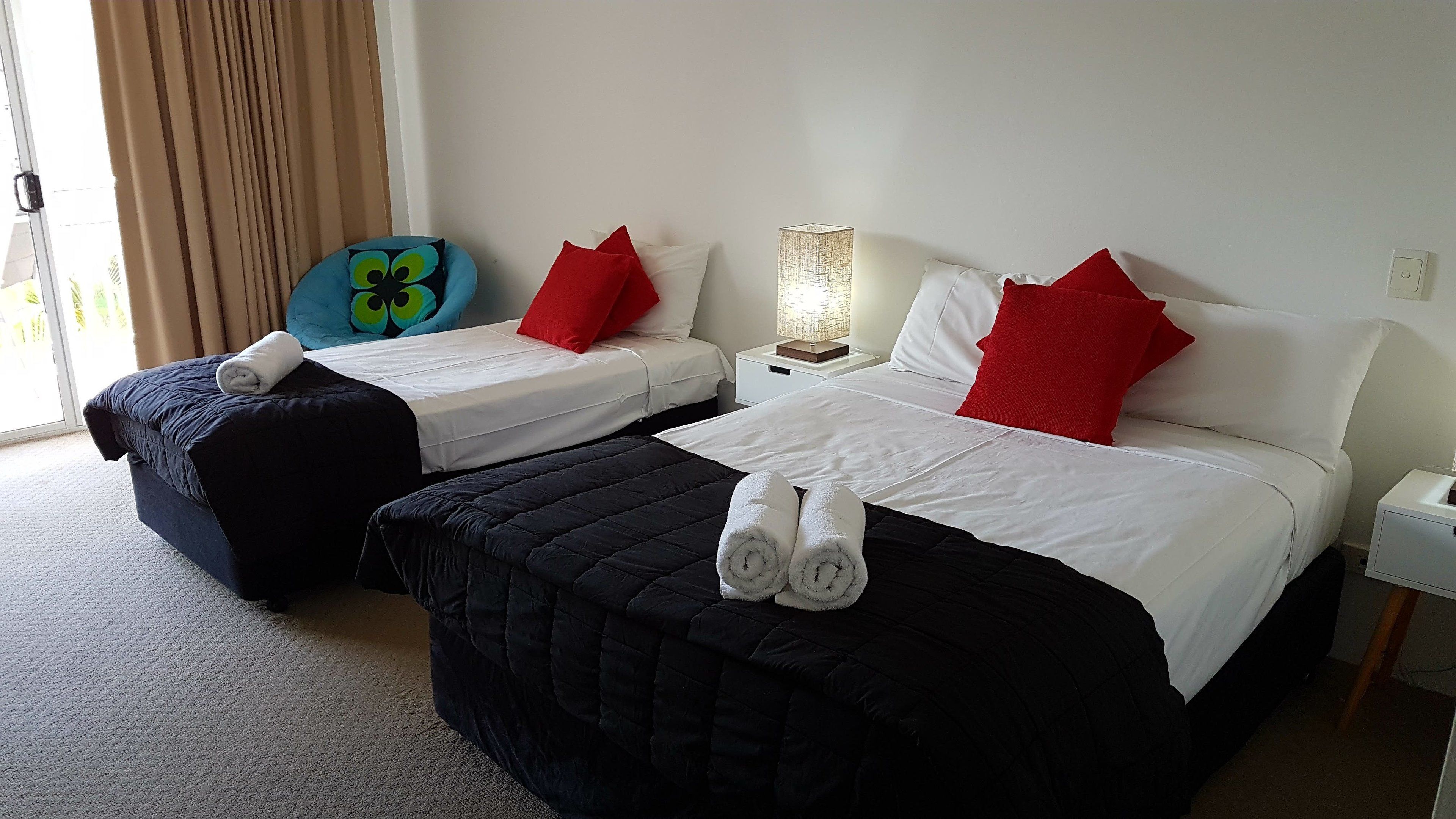 Habitació d'hotel amb dos llits, cadascú amb roba de llit blanca i almoines vermelles. Hi ha una almoina amb forma d'úvol blau a un dels llits, una làmpada a la tauleta de nit i una finestra amb cortines.