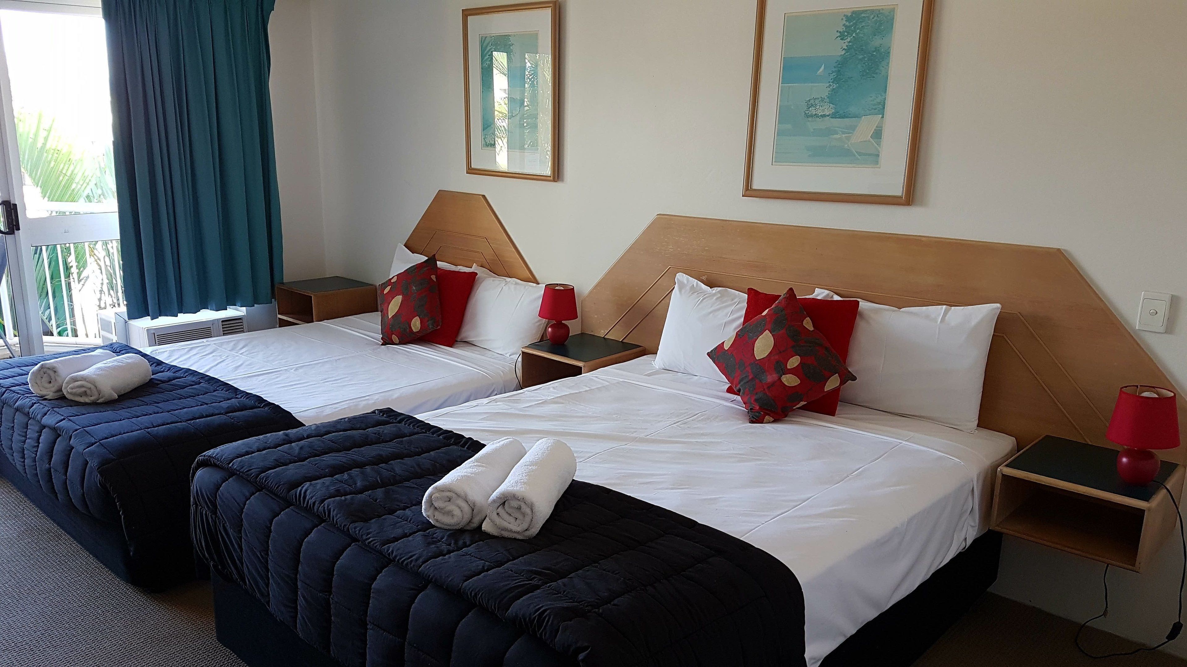 Une chambre d’hôtel avec deux lits, chacun doté d’une tête de lit, de draps blancs et de lampes rouges sur les tables de chevet. Deux tableaux encadrés ornent le mur, et il y a une fenêtre avec des rideaux.