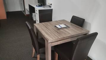 A small, neatly arranged room with a wooden table, four black chairs, a white desk with a computer and a printer, and a carpeted floor.