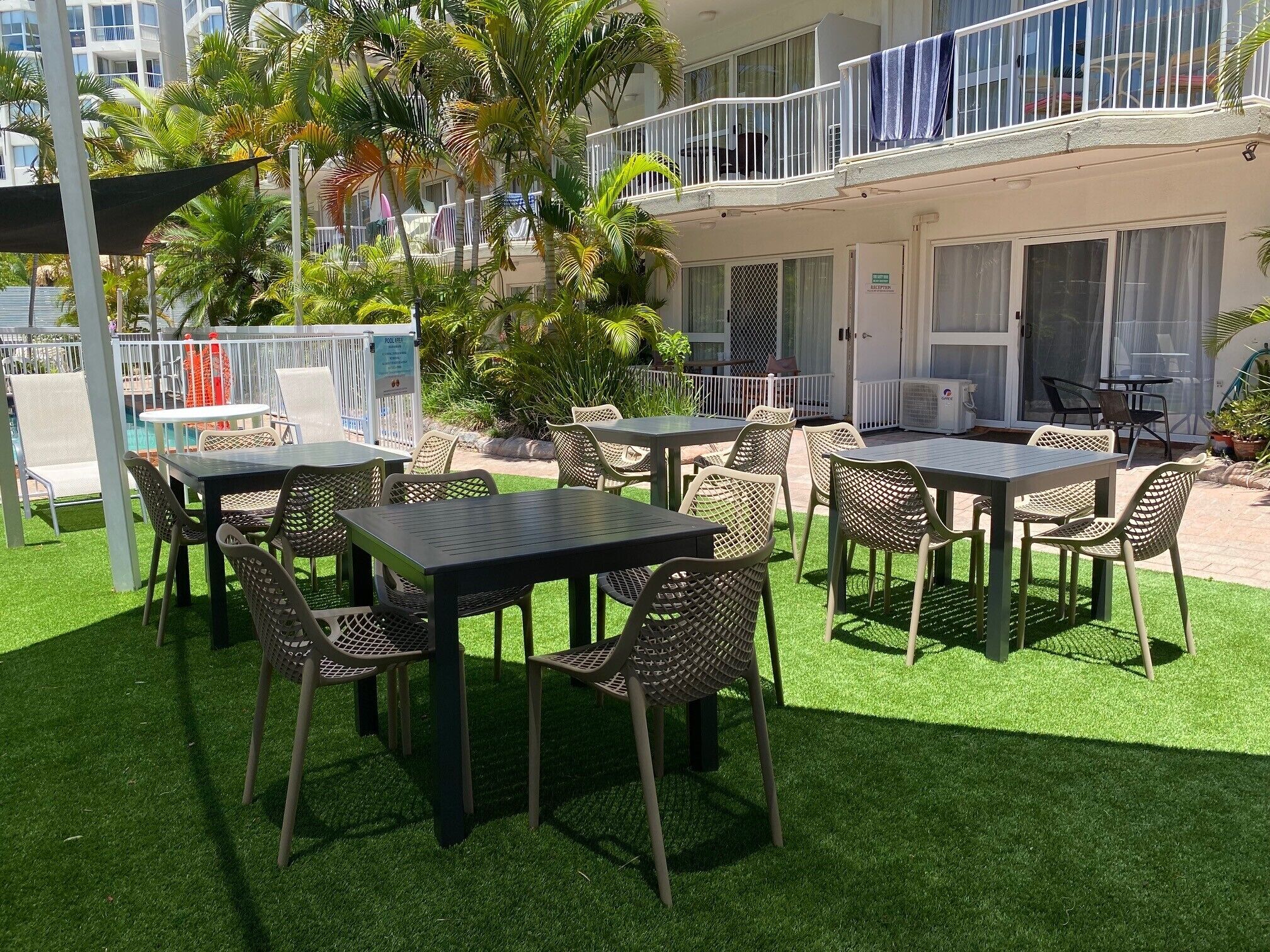An outdoor patio area with tables and chairs, surrounded by artificial grass, palm trees, and a pool.