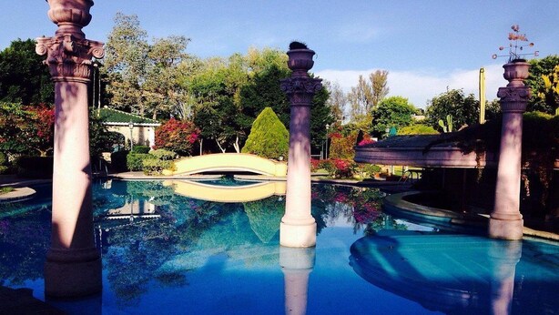 Una piscina con columnas clásicas y un puente, rodeada de exuberante vegetación y un cielo despejado.