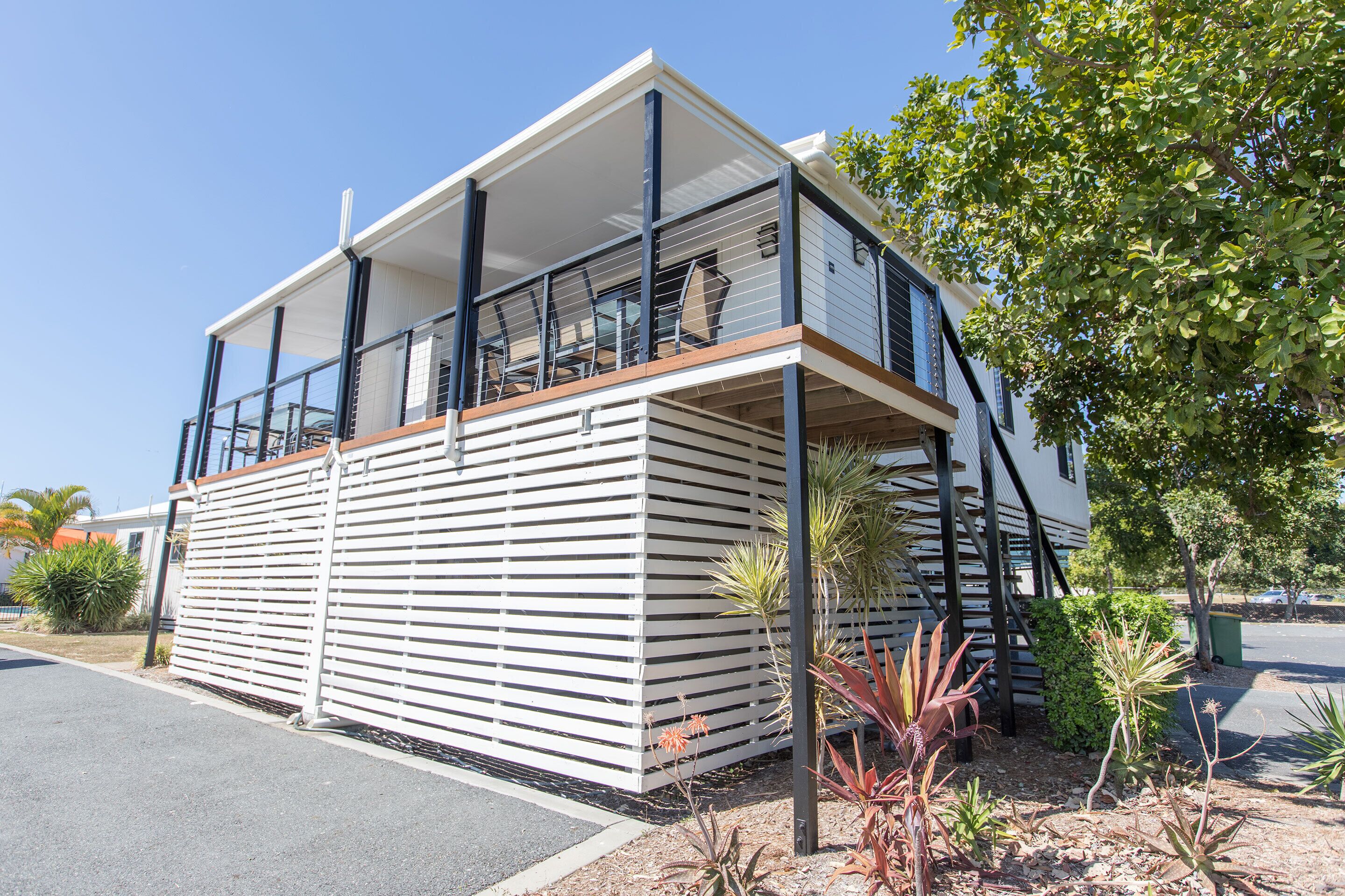 A modern two-story building with a balcony, surrounded by various plants and a clear sky.