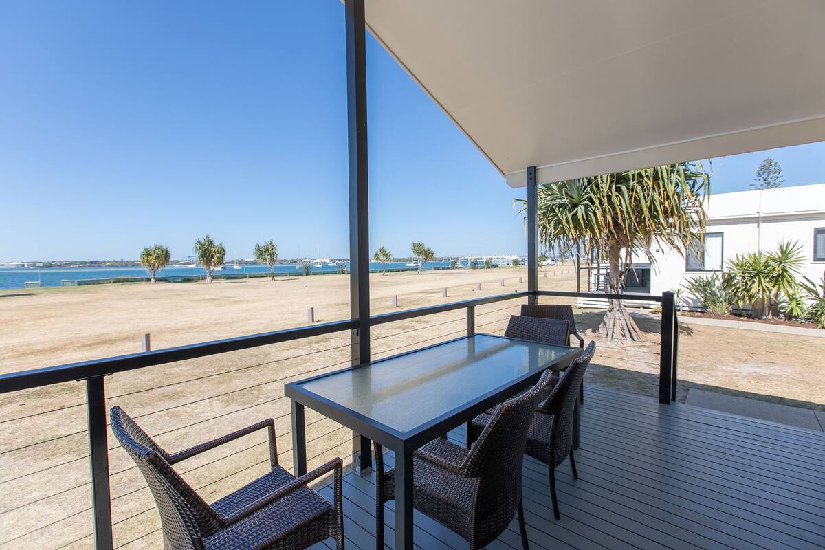 A balcony with a glass table and wicker chairs, overlooking a beach with palm trees and a clear blue sky.