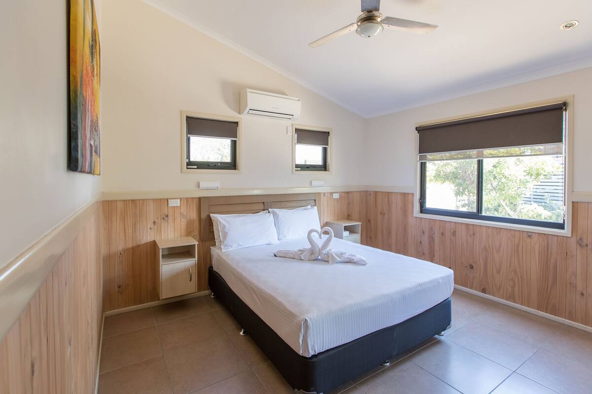 A bedroom with a bed, bedside tables, an air conditioning unit, a ceiling fan, and a window with blinds.