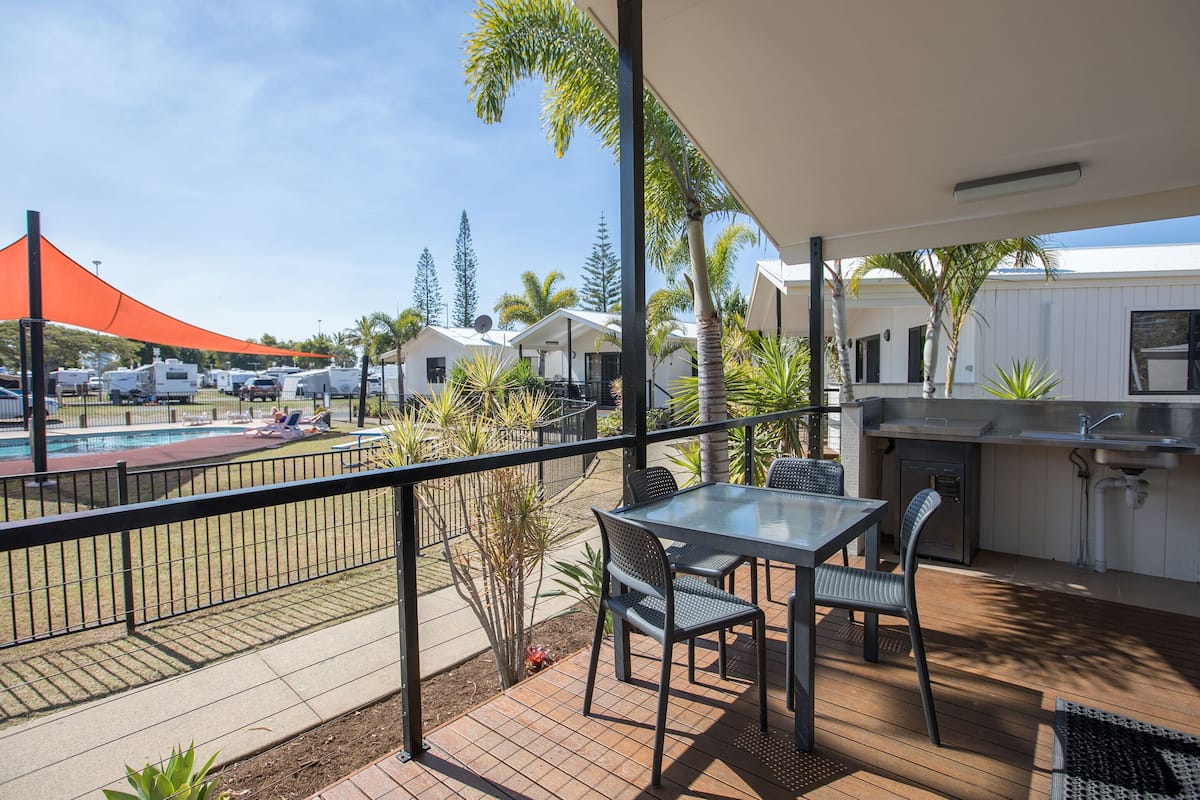 A balcony with a table and chairs, a barbecue area, and a view of a pool and RVs in the background.