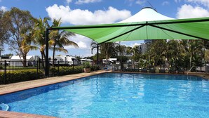 A swimming pool with a green canopy, surrounded by palm trees and a fence. - Broadwater Tourist Park (Southport)