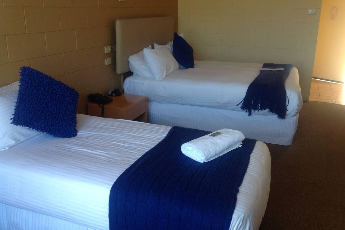 A hotel room with two beds, each with white linens and blue pillows. There is a wooden nightstand with a phone and a lamp between the beds.