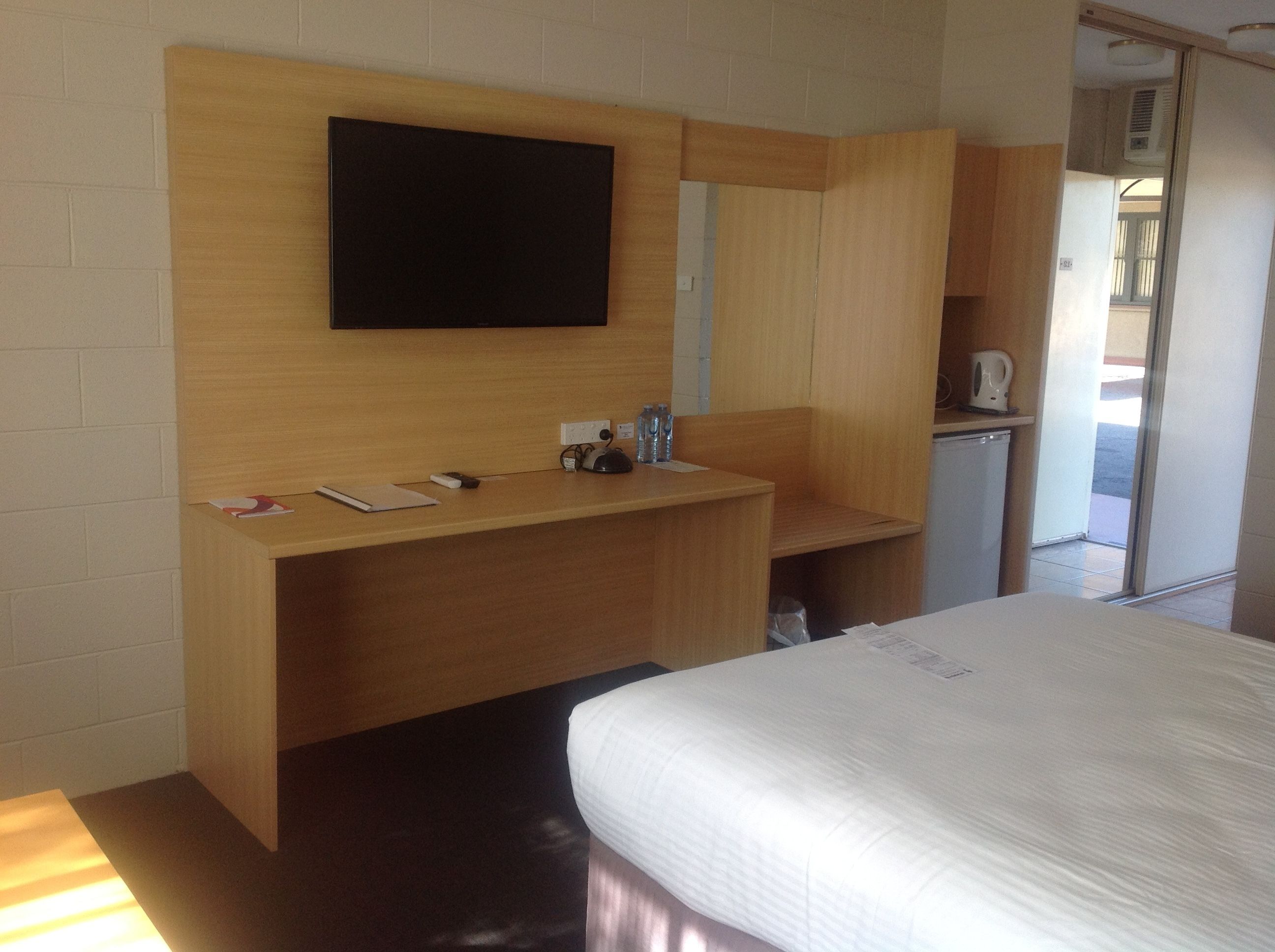 A hotel room with a wooden desk, a flat-screen TV mounted on the wall, a bed with white bedding, and a small kitchenette area.