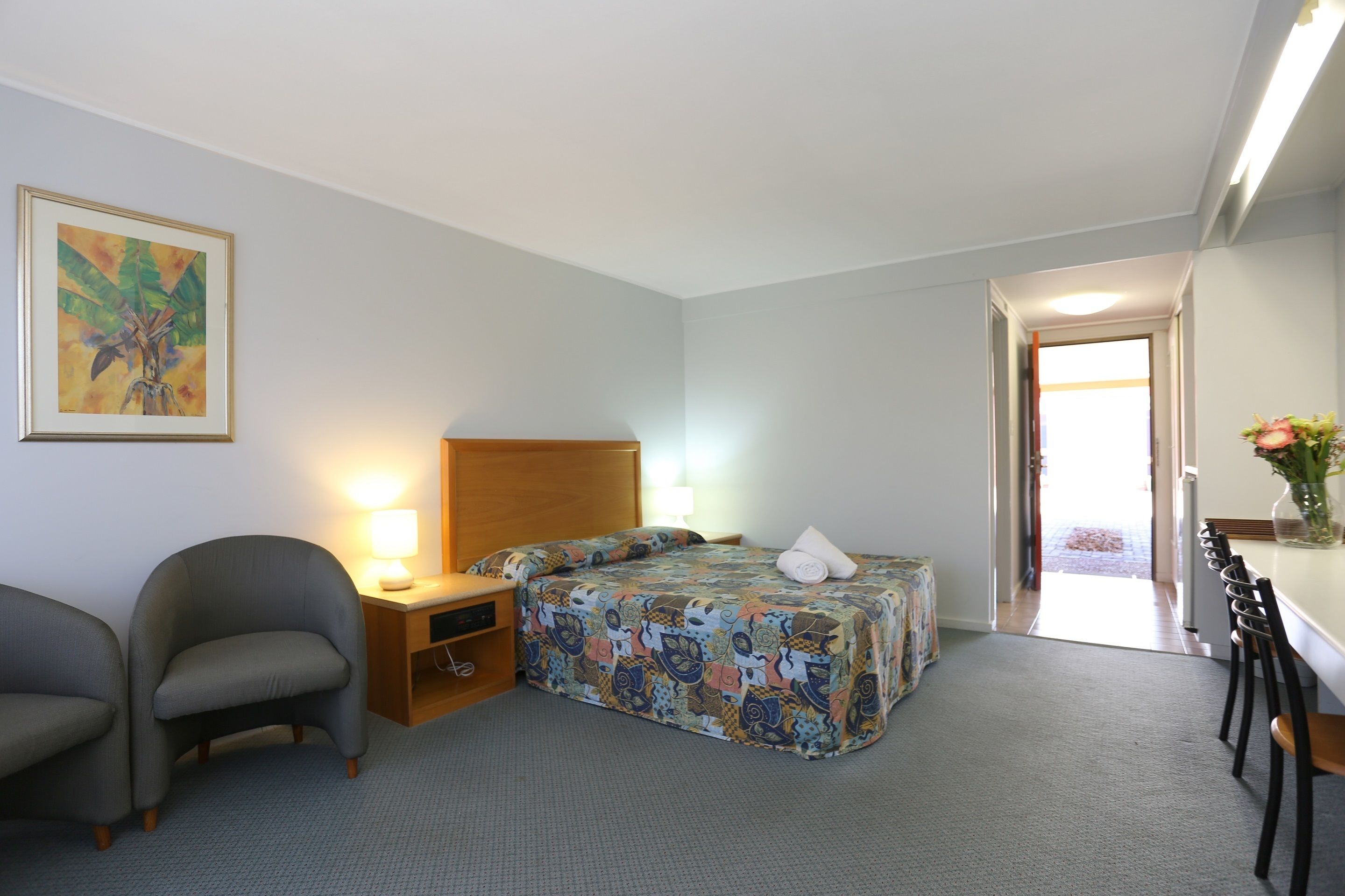 A hotel room with a bed, a desk, a chair, a nightstand, and a painting on the wall.