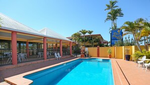 Piscina com área coberta e espaço para assentos ao ar livre, cercada por palmeiras e um edifício com a placa 'Reef Resort'.